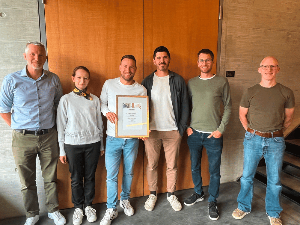 {
"alt": "Gruppenfoto vor einer Holztür mit Zertifikat.",
"title": "Gruppenfoto mit Zertifikat",
"caption": "Sechs Personen posieren mit einem Zertifikat vor einer Tür.",
"description": "Auf diesem Gruppenfoto halten sechs Personen ein Zertifikat in der Hand. Sie stehen lächelnd vor einer Holztür in einem Büro.",
}